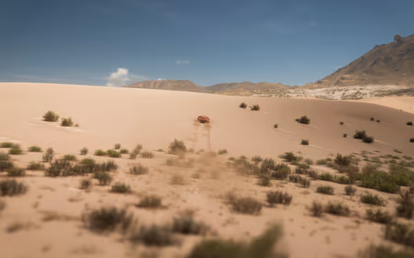 "Orange car driving across sandy desert dunes with scattered shrubs and mountains in the distance.