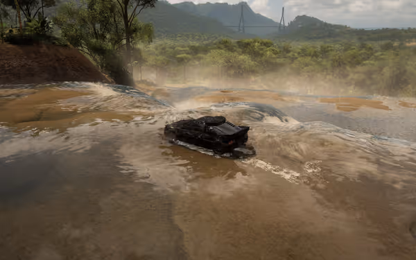 A black car crossing a shallow river surrounded by trees, hills, and a bridge.