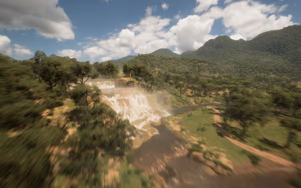 A large waterfall cascades through a green forest landscape with distant mountains under clouds.