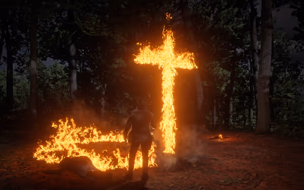 A male character stands before a burning cross in a forest at night.