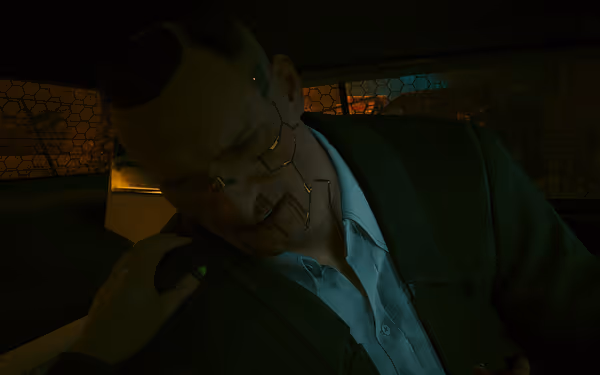 A cybernetic man with bloodied lips slumps in a car, a hand on his shoulder