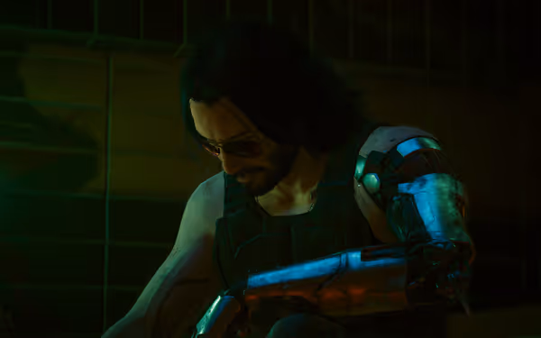A man with long hair, sunglasses, and a cybernetic arm sits in dim lighting.