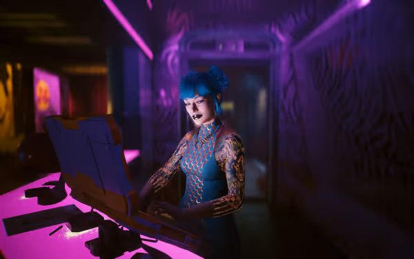 A woman with blue hair and tattoos types on a futuristic laptop in neon lighting.