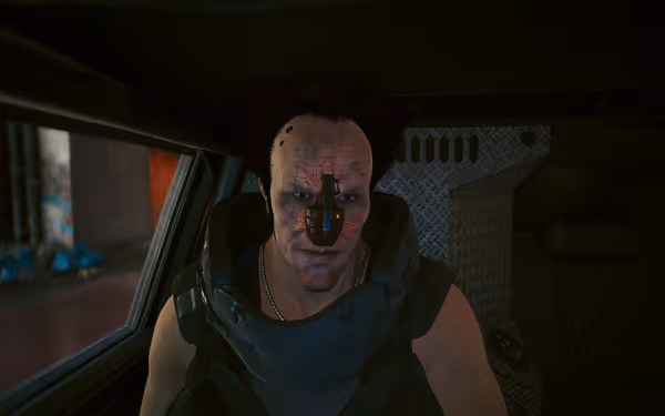 A muscular man with wild hair sits in a vehicle with a grenade embedded as his nose.