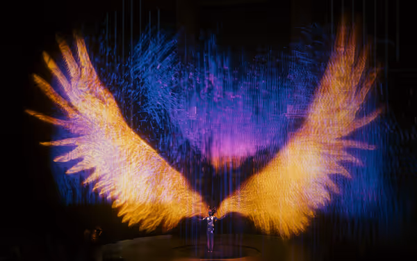 A performer stands beneath massive glowing orange and purple holographic wings on stage.