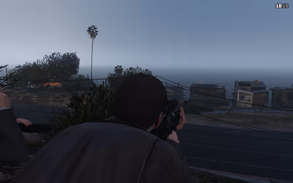 A man aims a rifle while lying on a hill, observing buildings by the ocean.