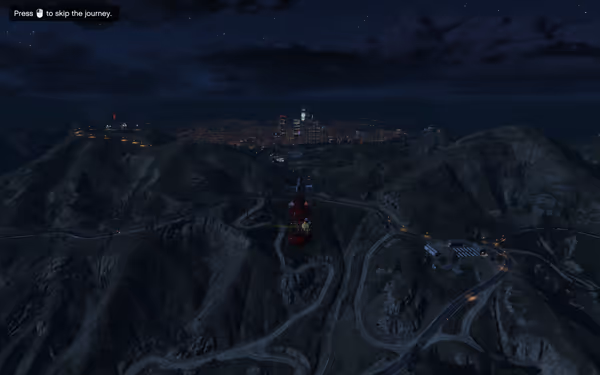 A red helicopter flies over winding mountain roads at night with a city glowing in the distance.