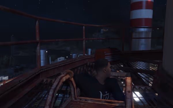 A person sits on a rusted platform at night, overlooking distant lights and structures.