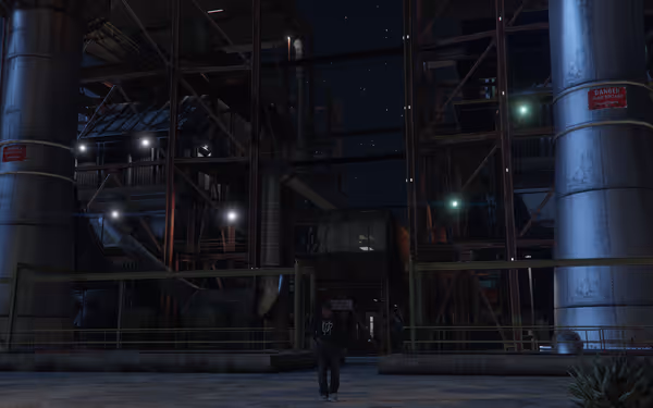 A person in casual clothing stands in an industrial area at night, with large pipes and warning signs around.