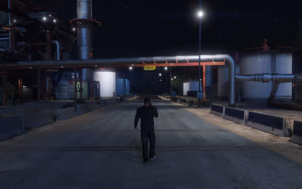 A person walks down an industrial road at night, passing large tanks and pipes with warning signs.