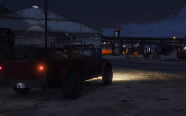 A red off-road vehicle with its lights on is parked near warehouses and industrial structures.