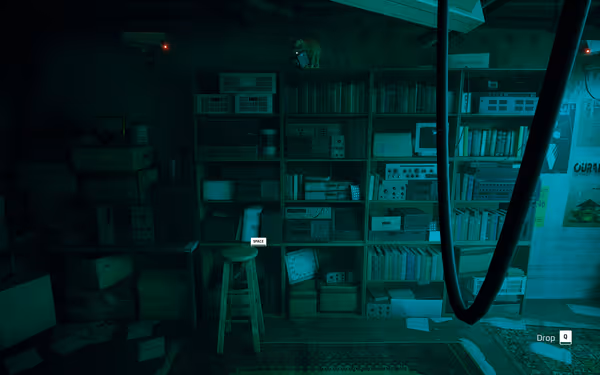 A cat sits on top of a bookshelf in a dimly lit, cluttered room with electronics.